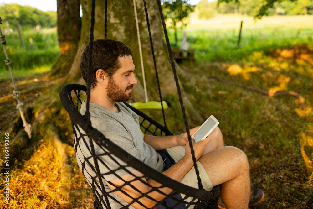 Photo of a young and attractive guy sitting on a hammock relaxed reading an electronic book. It is a summer day in the countryside of Scotland