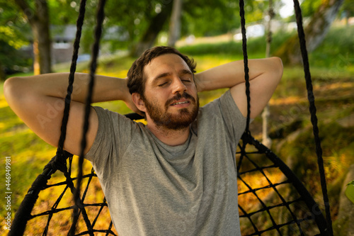Photo of a attractive young guy relaxing on a hammock in the middle of nature. It is a summer day in the forest and he has a happy smile