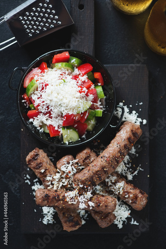 Close-up of grilled cevapi or cevapcici with serbian salad on a black wooden serving board, vertical shot, top view