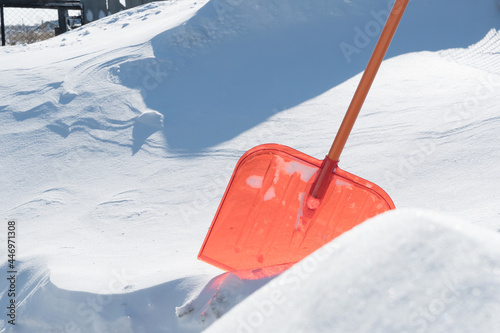 A snow shovel is stuck in a snowdrift in winter in winter. Snow removal, cleaning the street from snow drifts. A white pile of snow. Snowy frost weather in winter. A red shovel. Snowy Crystals © Yevgeniy