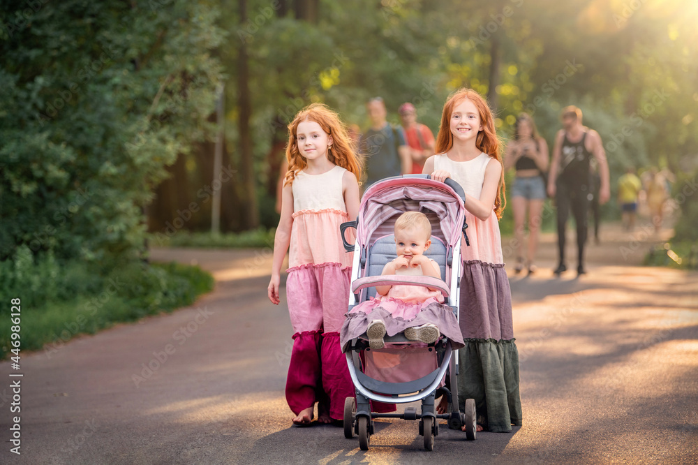 Two older redhead sisters are walking in park on a sunny summer evening. with a stroller with her little sister. Girls in long vintage dresses roll stroller along the path Two older redhead sisters are walking in park on a sunny summer evening. with a stroller with her little sister. Girls in long vintage dresses roll stroller along the path
