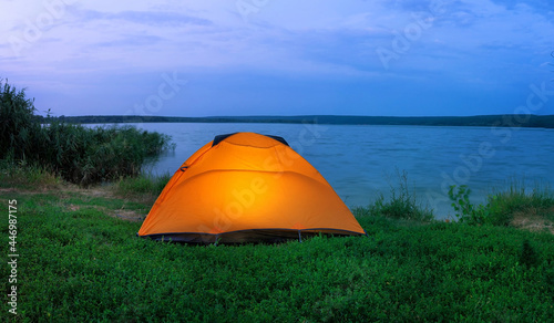 Orange tent illuminated from the inside by lake at dusk