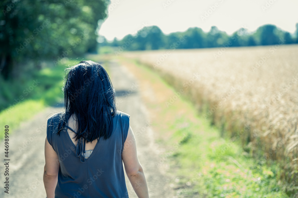 Young woman with black hair is walking along a dirt road between forest and wheat field in sunny weather Young woman with black hair is walking along a dirt road between forest and wheat field in sunny weather