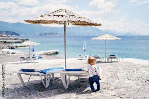 Child stands near a sun lounger on the beach and looks to the side. Back view