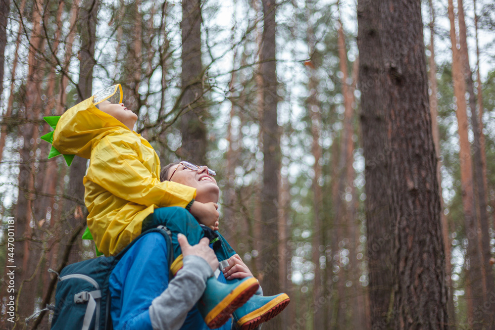 Mom and child walking in the forest after rain in raincoats together, the child is sitting on the shoulders of mom, happily looking at the sky Mom and child walking in the forest after rain in raincoats together, the child is sitting on the shoulders of mom, happily looking at the sky