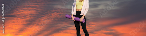 A sporty slim girl in leggings and a top is standing with a training mat, resting between exercises. Fitness and healthy lifestyle.