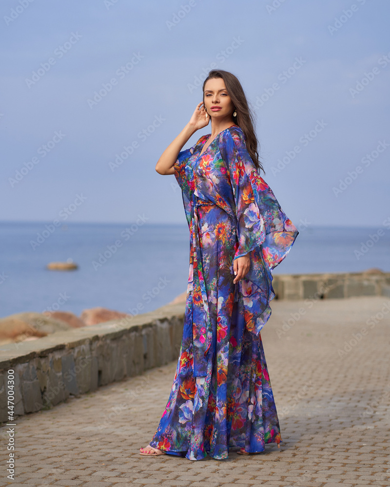 Elegant caucasian woman with wavy brunette hair in vivid blue colorful dress walking outdoors on a sunny summer day at pavement against view on s sea water Elegant caucasian woman with wavy brunette hair in vivid blue colorful dress walking outdoors on a sunny summer day at pavement against view on s sea water