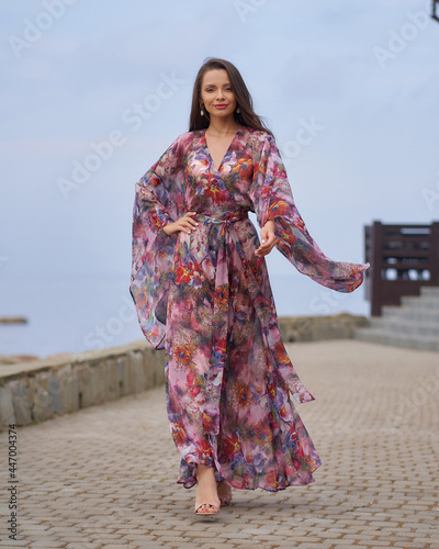 Young fashionable woman in colorful pnk dress with floral design walking at city embanlment with pavement against sea and sky. photo