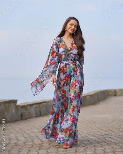 Young beautiful and elegant woman with long brunette hair wearing summer dress with floral design and posing against sea and sky