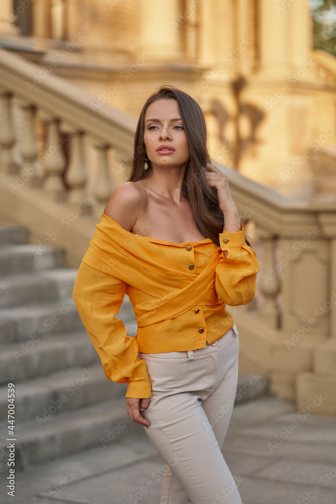 Fototapeta premium Elegant stylish fashionable woman with long wavy hair posing in orange top against stone stairs in the city on a summer sunny evening