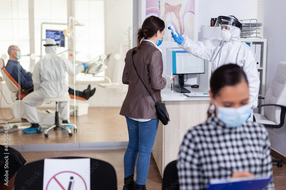 Naklejka premium Stomatology receptionist using digital thermometer to measure patient temperature dressed in ppe suit as safety precaution during global outreabk with coronavirus. Dentist treating teeth.