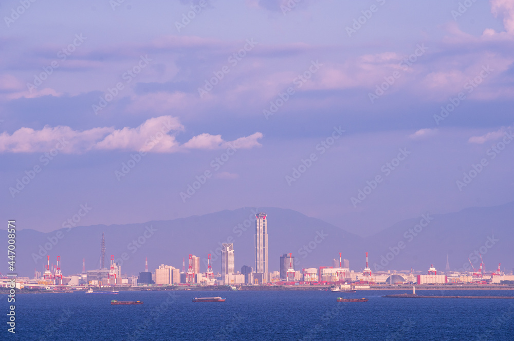 夏の夕暮れ、大阪湾に入道雲が浮かぶ。夕陽がさして雲が少しオレンジ色に染まる。海の向こう岸、遠くには大阪の南港と高層ビル群が見える。神戸からの対岸の眺め。