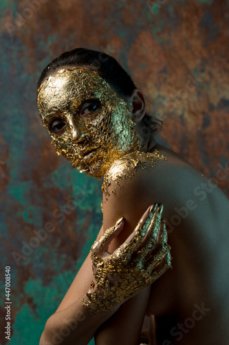 Girl with a mask on her face made of gold leaf. Gloomy studio portrait of a brunette on an abstract background.