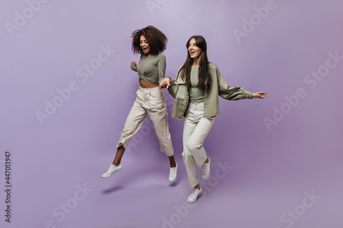 Wavy haired lady in long sleeve top and cool pants jumping and holding hands with modern woman in olive and beige clothes.