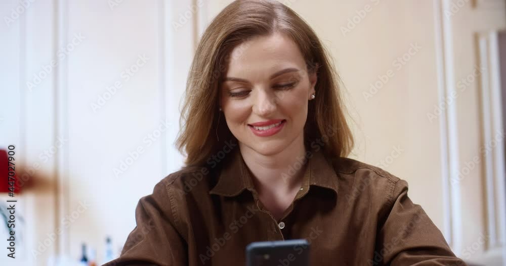 Online chat. Smiling woman talking on smartphone with friends while sitting at the office. Portrait of a positive girl sitting indoors and typing at the phone while having pleasure conversation with Online chat. Smiling woman talking on smartphone with friends while sitting at the office. Portrait of a positive girl sitting indoors and typing at the phone while having pleasure conversation with