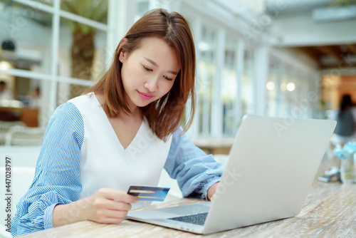 Portrait A beautiful Asian woman in a white shirt sitting in a bakery shopping online with her credit card and laptop computer in front of her hands.