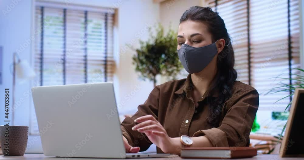 Portrait positive cheerful beautiful girl wearing protective mask typing at the keyboard while reading news or messages at the laptop. Satisfied woman sitting at her workplace at office. Job concept Portrait positive cheerful beautiful girl wearing protective mask typing at the keyboard while reading news or messages at the laptop. Satisfied woman sitting at her workplace at office. Job concept
