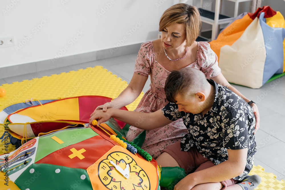 Children with disability getting sensory activity with toys, balls, small objects, cerebral palsy boy playing calming game, training fine motor skills. Rehabilitation center with therapist, mother - obrazy, fototapety, plakaty 