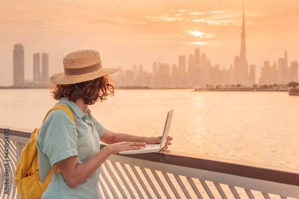 Woman blogger or copywriter or student works and studies using a laptop and admires the colorful sunset over Dubai Creek with a view of the skyscrapers and the Burj Khalifa