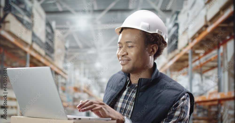 Young African-American warehouse worker standing on backround of row with load and communicates over video call using laptop. Young African-American warehouse worker standing on backround of row with load and communicates over video call using laptop.