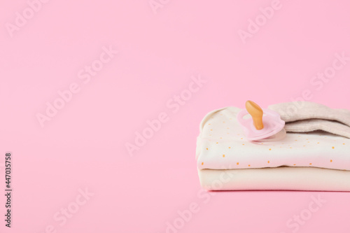 Stack of baby clothes and pacifier on color background