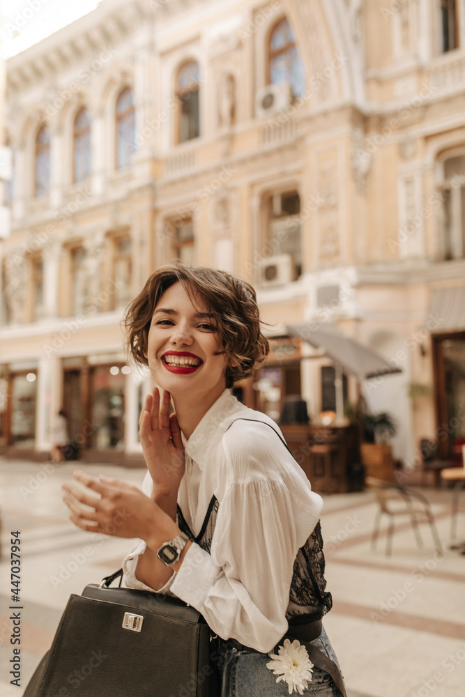 Positive girl with bright red lips and wavy hair laughing in city. Cool woman in white shirt with handbag posing at street..