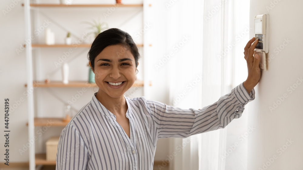 Head shot portrait of smiling Indian woman using smart home system controller on wall, happy young female looking at camera, switching temperature on thermostat or activating or turning off alarm Head shot portrait of smiling Indian woman using smart home system controller on wall, happy young female looking at camera, switching temperature on thermostat or activating or turning off alarm