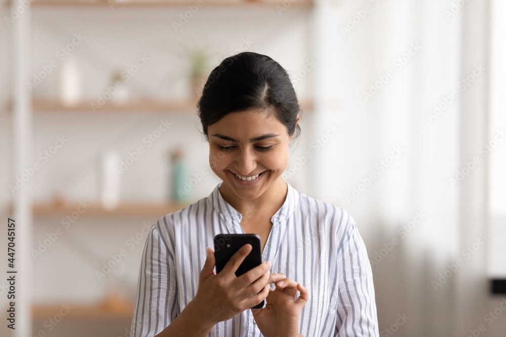 Fototapeta premium Head shot smiling Indian woman using smartphone, having fun with device, standing at home, positive attractive young female typing writing message in social network, chatting or shopping online