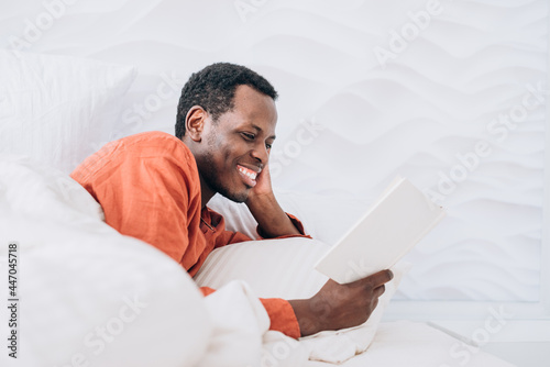 Positive African-American man in pajamas reads interesting novel lying in comfortable bed under soft blanket in room closeup