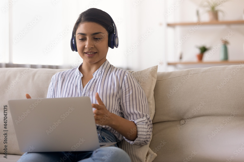 Naklejka premium Indian woman in headphones chatting online in social network, using laptop, sitting on couch at home, young female student freelancer businesswoman involved in internet meeting, making video call