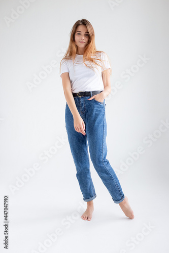 young attractive caucasian woman with long hair in t-shirt and blue jeans isolated on white studio background. skinny pretty female posing on cyclorama with bare feet. model tests of beautiful lady