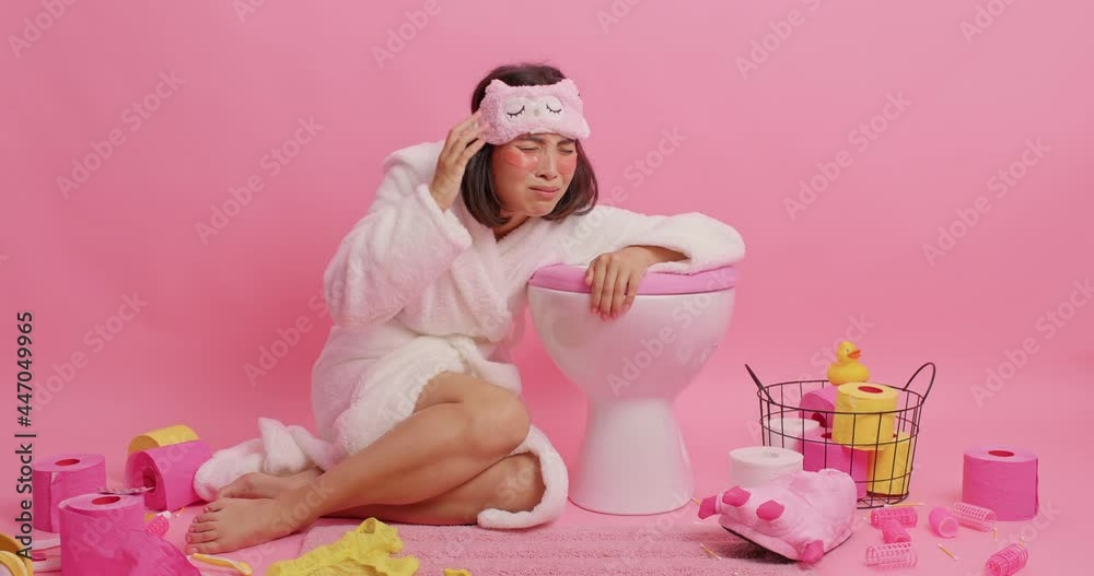 Unhappy young Asian woman feels unwell after hangover suffers from nausea and stomachache leans on toilet bowl wears sleepmask white bathrobe spends all night in lavatory room takes painkillers Unhappy young Asian woman feels unwell after hangover suffers from nausea and stomachache leans on toilet bowl wears sleepmask white bathrobe spends all night in lavatory room takes painkillers