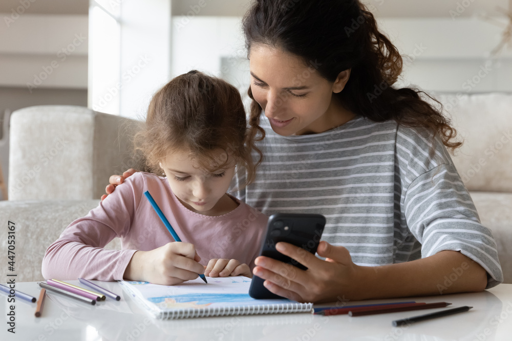 Loving young Latino mother and teen daughter draw paint with cellphone online lesson or class. Caring Hispanic mom and small girl child engaged in online education activity on smartphone together. Loving young Latino mother and teen daughter draw paint with cellphone online lesson or class. Caring Hispanic mom and small girl child engaged in online education activity on smartphone together.