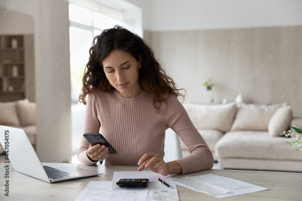 Young Hispanic woman sit at desk at home manage household finances paying bills calculating on machine cellphone. Millennial Latin female care of budget use smartphone for payment. Finance concept. - obrazy, fototapety, plakaty 