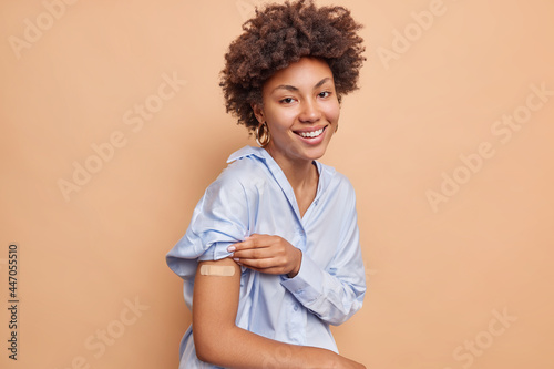 Pretty pleased Afr American woman in blue shirt raises sleeve shows vaccinated arm wears adhesive plaster smiles pleasantly isolated over beige background. Covid 19 protection and immunization photo