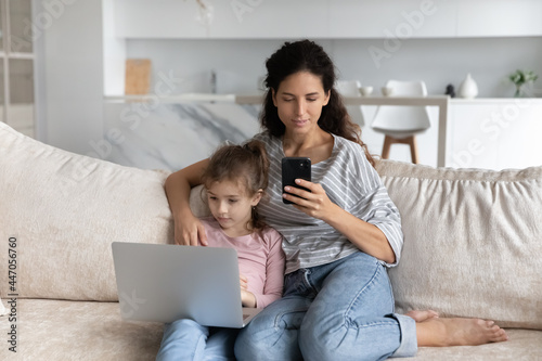 Young Hispanic mother and teen biracial daughter sit rest on sofa at home use electronic gadgets together. Latin mom and small girl child relax in living room browsing devices. Technology concept.