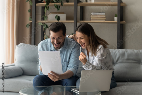 Young 30s couple feels euphoric look very happy scream with joy read good unbelievable news in paper letter document. Overjoyed spouses get pleasant message celebrate opportunity receive offer concept photo