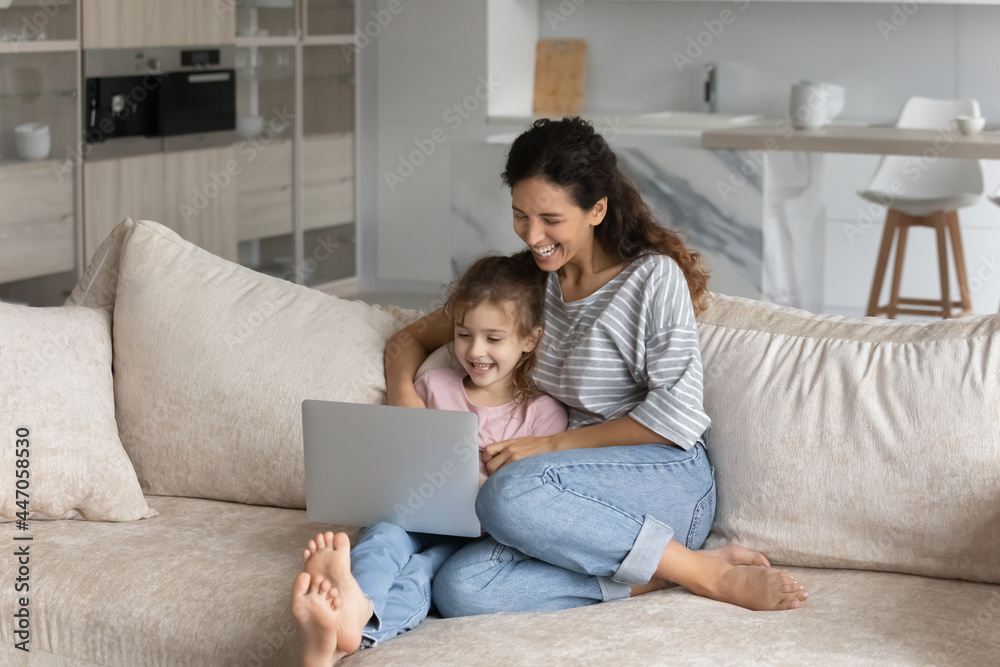 Smiling Latino mom and small teen daughter relax on couch at home use modern computer gadget together. Happy Hispanic mother and teenage girl child rest on sofa in living room have fun browse laptop.