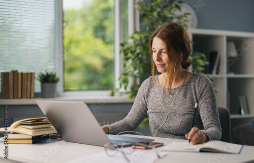 Beautiful woman in earphones sitting at living room and using modern laptop for for online education. Focused female taking notes while studying at home.