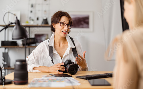 Professional photographer sitting at bright office and talking with client about photo session.Two women discussing working details indoors.
