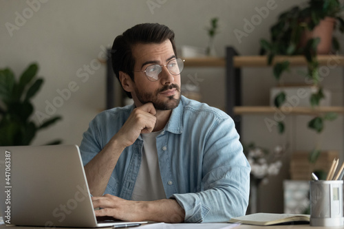 Thoughtful young business man in glasses sit at desk distracted from computer work looks into distance consider contemplate search solution or fresh creative startup ideas. Brooding, brainwork concept