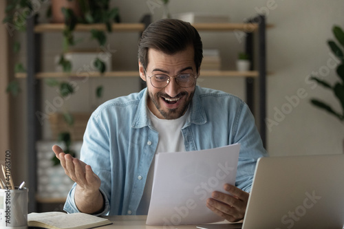 Surprised guy read exciting news feels happy got job promotion letter looks shocked. Man in glasses sit at homeoffice desk hold document papers sheets excited by money refund, great sale offer concept