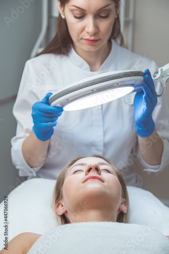 Beautician doctor is examining woman's face with special lamp