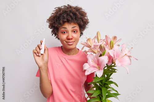 Obraz na plátne Indoor shot of curly haired woman holds nasal spray found proper medicament for stuff nose suffers from allergic rhinitis holds lilies reacts on pollen has red watery eyes