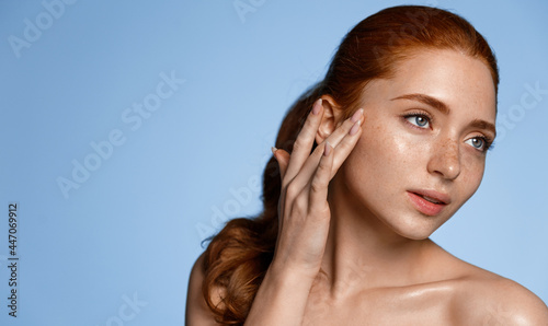 Women wellbeing. Attractive redhead girl with freckles, touching smooth, perfect clean skin without makeup, gently touch glowing healthy face and gazing aside, skincare concept, blue background photo