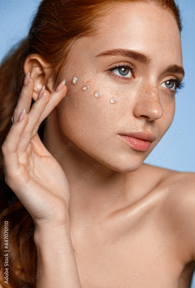 Close up of beautiful redhead woman, nude no makeup, using facial skin care  cream, rub in cosmetic product on face for glowing, healthy and young face  effect, blue background Stock Photo