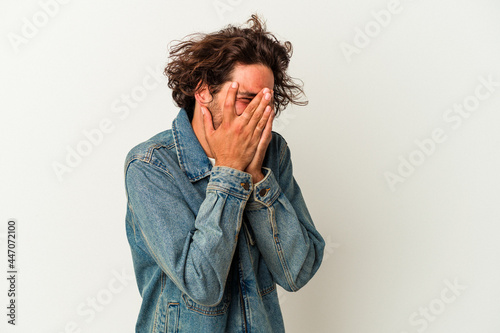 Young caucasian man isolated on white background blink at the camera through fingers, embarrassed covering face.