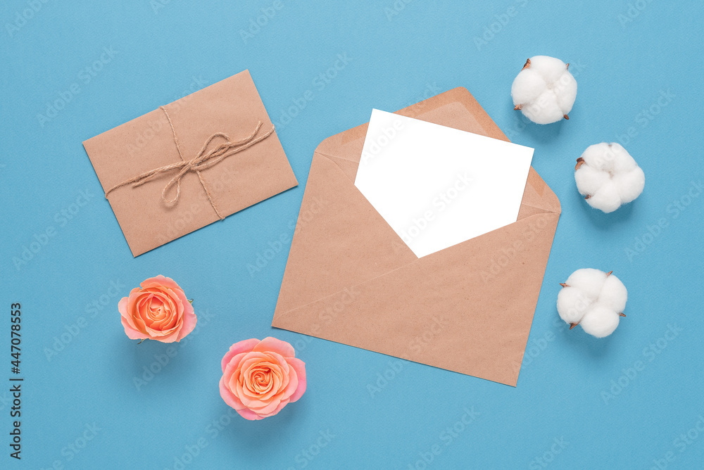 Blank card mockup in an envelope on a blue paper background decorated with roses, cotton flowers. Wedding stationery template. Top view, flat lay. Blank card mockup in an envelope on a blue paper background decorated with roses, cotton flowers. Wedding stationery template. Top view, flat lay.