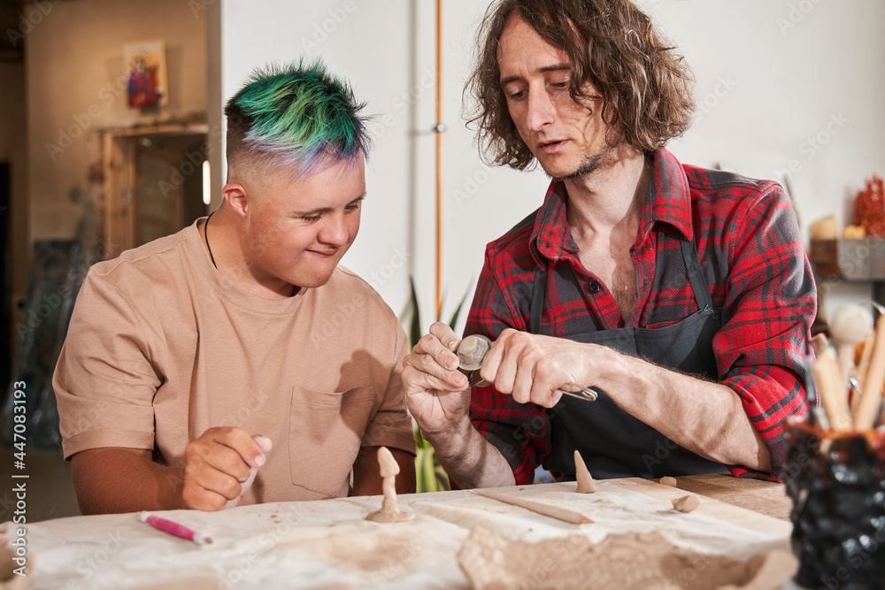 Craftsman showing to his student with down syndrome how to sculpting from the clay - obrazy, fototapety, plakaty 