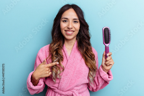 Young mexican woman wearing a bathrobe holding a brush isolated on blue background person pointing by hand to a shirt copy space, proud and confident